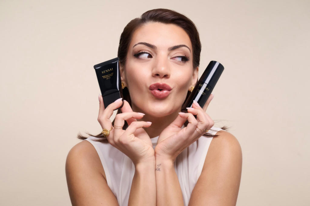 Katia Mosally in a white top and red lips, holdig one SENSAI product in each hand.