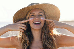 A girl, wearing a sun hat, applies sunscreen to her cheeks.